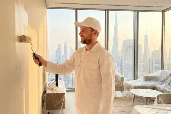 A skilled painter from Dubai Technical Works applying smooth beige emulsion paint to a modern apartment wall, with sunlight streaming through floor-to-ceiling windows overlooking the Dubai skyline